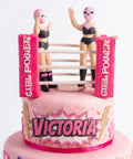 Detalle del ring con dos luchadoras modeladas a mano y postes Girl Power sobre pastel de fondant rosa, L'Autrichienne CDMX