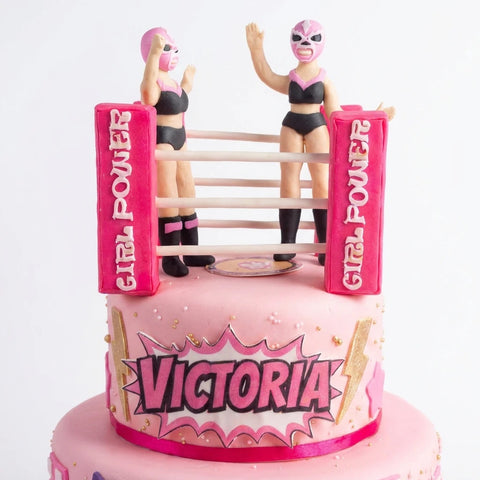 Detalle del ring con dos luchadoras modeladas a mano y postes Girl Power sobre pastel de fondant rosa, L'Autrichienne CDMX
