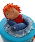 Detalle de la figura de Ponyo modelada a mano con cabello naranja y vestido rojo entre peces azules sobre una ola de fondant, L'Autrichienne CDMX