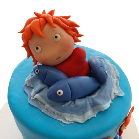Detalle de la figura de Ponyo modelada a mano con cabello naranja y vestido rojo entre peces azules sobre una ola de fondant, L'Autrichienne CDMX